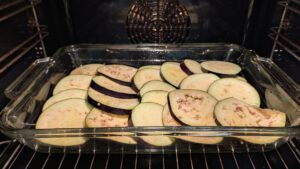 Rohe Auberginenscheiben mit violetter Schale liegen überlappend in einer rechteckigen Glasform im Ofen; Kartoffelscheiben sind nicht zu sehen.
