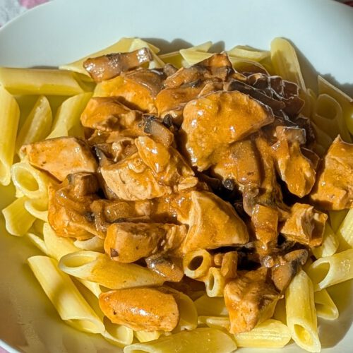 Cremiges Chicken Stroganoff mit Champignons auf glutenfreien Penne, auf einem Teller auf einer Tischdecke mit Herzmuster im Sonnenlicht