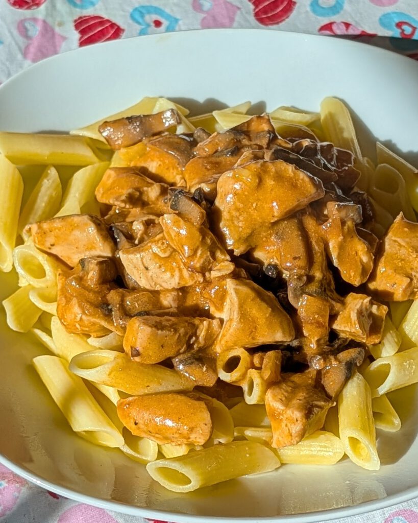 Cremiges Chicken Stroganoff mit Champignons auf glutenfreien Penne, auf einem Teller auf einer Tischdecke mit Herzmuster im Sonnenlicht