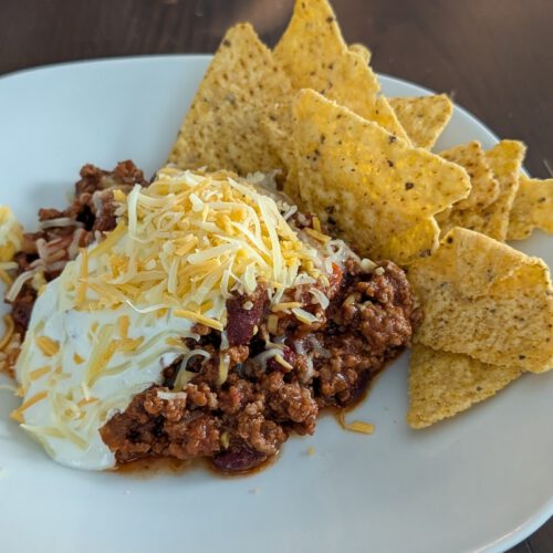 Weißer Teller mit Chili con Carne und Kidneybohnen, darauf Saure Sahne und geriebener Cheddar, rechts gelbe Tortilla-Chips, auf einem dunklen Holztisch