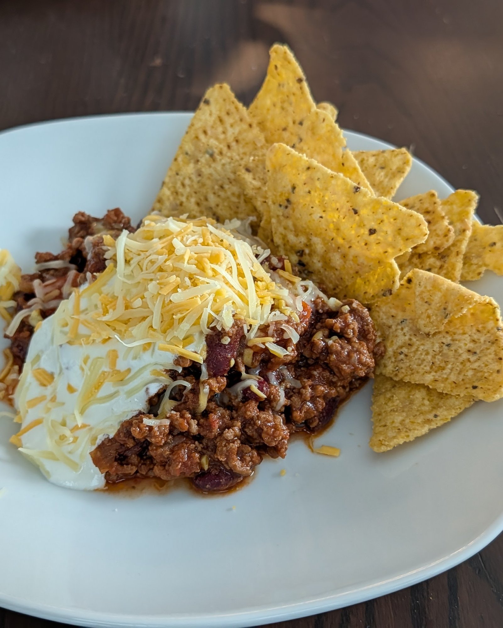 Weißer Teller mit Chili con Carne und Kidneybohnen, darauf Saure Sahne und geriebener Cheddar, rechts gelbe Tortilla-Chips, auf einem dunklen Holztisch