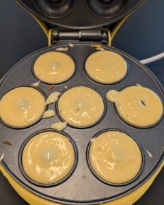 Donutteig in den sieben Mulden eines geöffneten gelben Donut-Makers, mit ein paar Tropfen auf der Platte