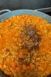 Pfanne mit orangefarbener Hackfleisch-Tomatensoße und roten Paprikastücken, in der Mitte ein Häufchen fein gehackte dunkle Schokolade vor dem Unterrühren
