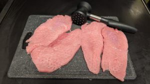 Vier rohe, rosa Schnitzel flach geklopft auf einem dunkelgrauen Schneidebrett, dahinter liegt ein schwarzer Fleischklopfer