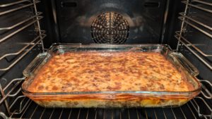 Gebackene Moussaka in einer rechteckigen Glasform im Ofen mit goldbrauner, dunkel gesprenkelter Oberseite; am Rand der Form sind Schichten zu erkennen.