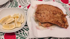 Zwei goldbraun gebackene Schnitzel auf Küchenpapier neben einer weißen Schale mit Zitronenspalten, auf einer rot-grün gemusterten Tischdecke