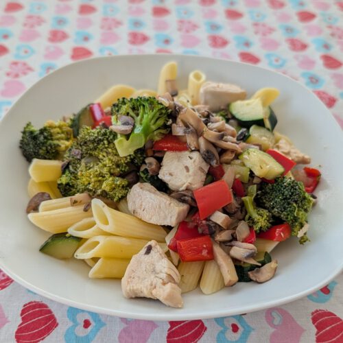 Weißer Teller mit Penne, Hähnchenstücken, Brokkoli, roter Paprika, Zucchini und Champignonscheiben auf einer Tischdecke mit Herzmuster