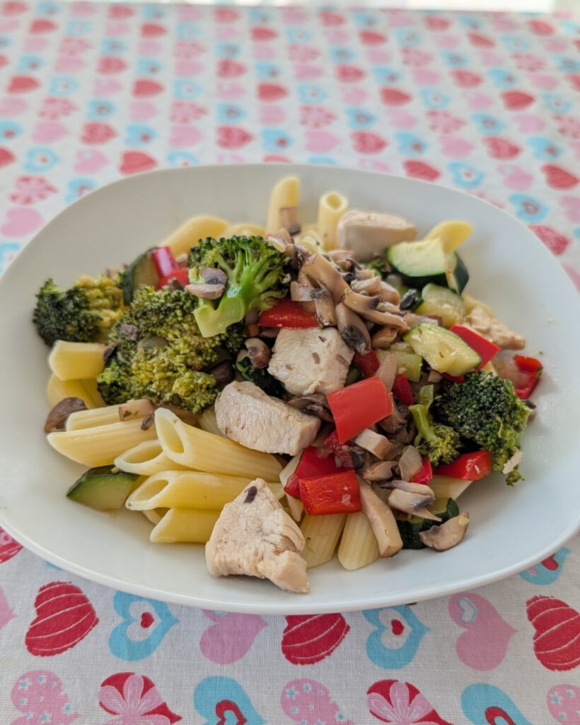 Weißer Teller mit Penne, Hähnchenstücken, Brokkoli, roter Paprika, Zucchini und Champignonscheiben auf einer Tischdecke mit Herzmuster