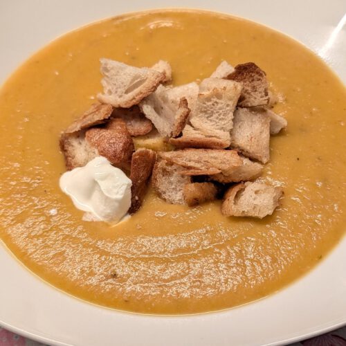 Weißer, eckiger Teller mit cremiger orangefarbener Karotten-Kartoffel-Suppe, goldenen Croutons und einem Klecks saurer Sahne auf einer Tischdecke mit Herzmuster.