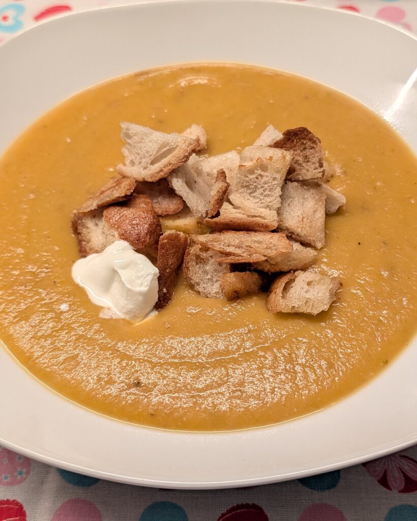 Weißer, eckiger Teller mit cremiger orangefarbener Karotten-Kartoffel-Suppe, goldenen Croutons und einem Klecks saurer Sahne auf einer Tischdecke mit Herzmuster.