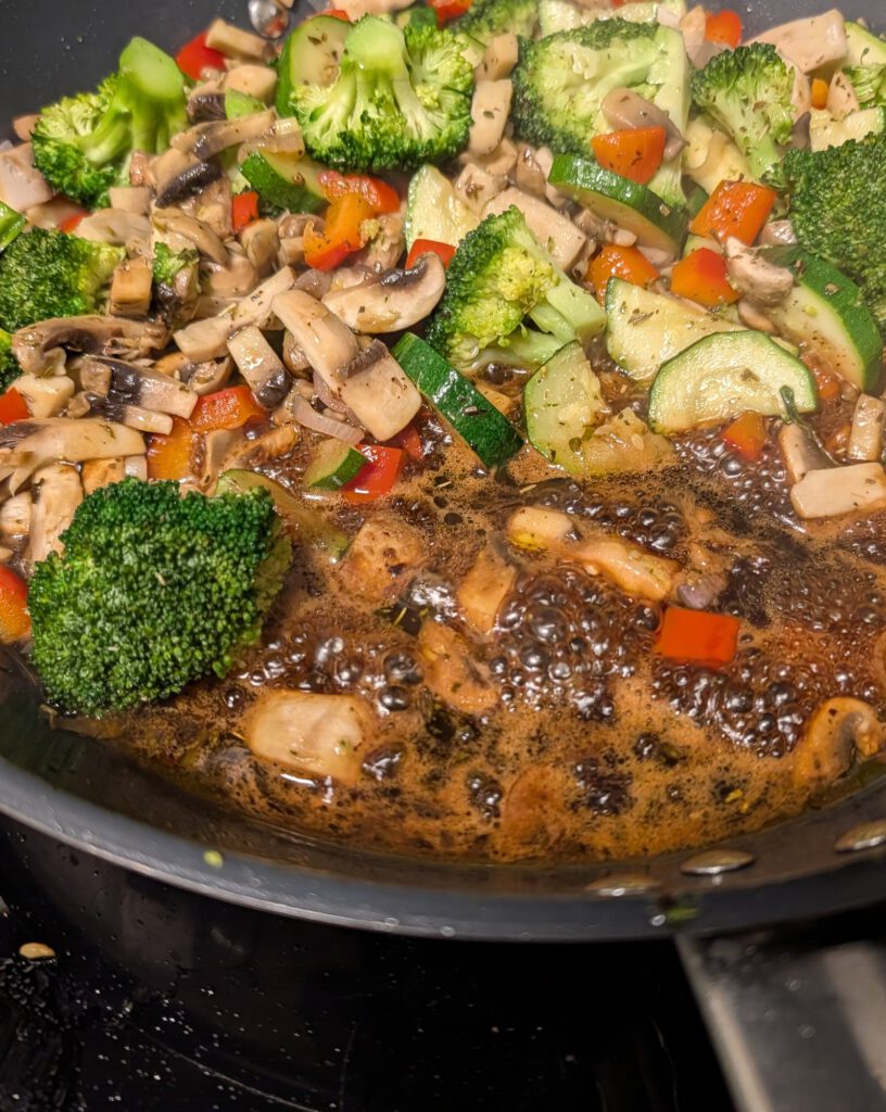 Pfanne voller Brokkoliröschen, Champignonscheiben, Zucchini und roter Paprika, unten blubbert dunkler Balsamico-Sirup.