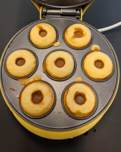 Sieben frisch gebackene goldgelbe Mini-Donuts in den geöffneten Mulden eines gelben elektrischen Donut-Makers, von oben fotografiert