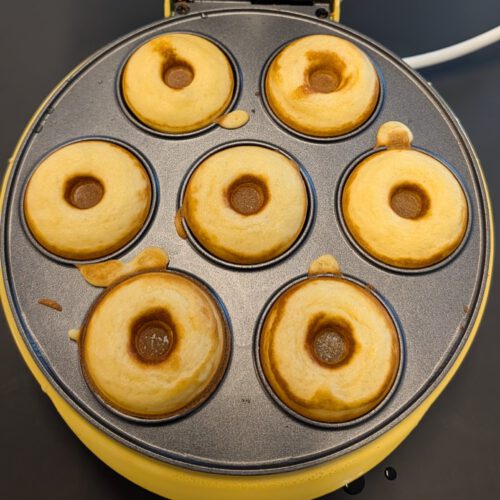 Sieben frisch gebackene goldgelbe Mini-Donuts in den geöffneten Mulden eines gelben elektrischen Donut-Makers, von oben fotografiert