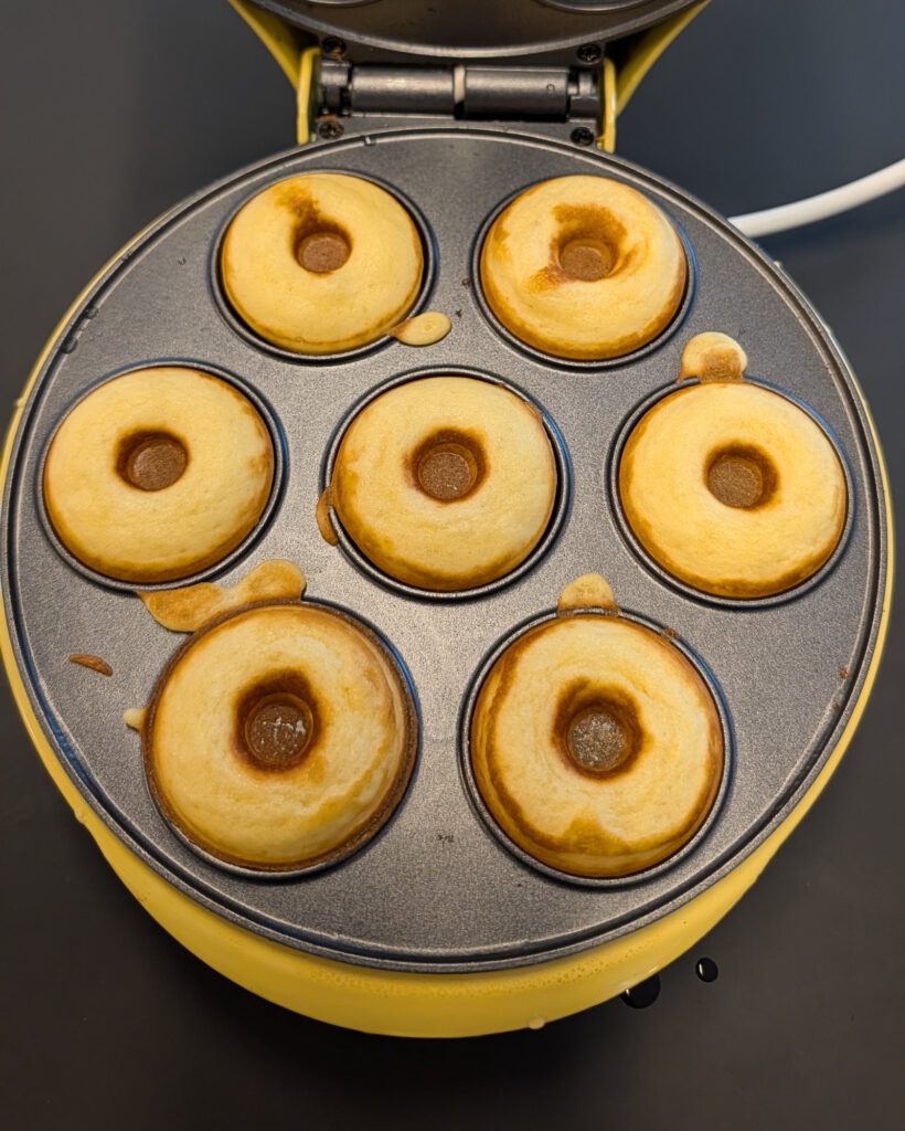 Sieben frisch gebackene goldgelbe Mini-Donuts in den geöffneten Mulden eines gelben elektrischen Donut-Makers, von oben fotografiert
