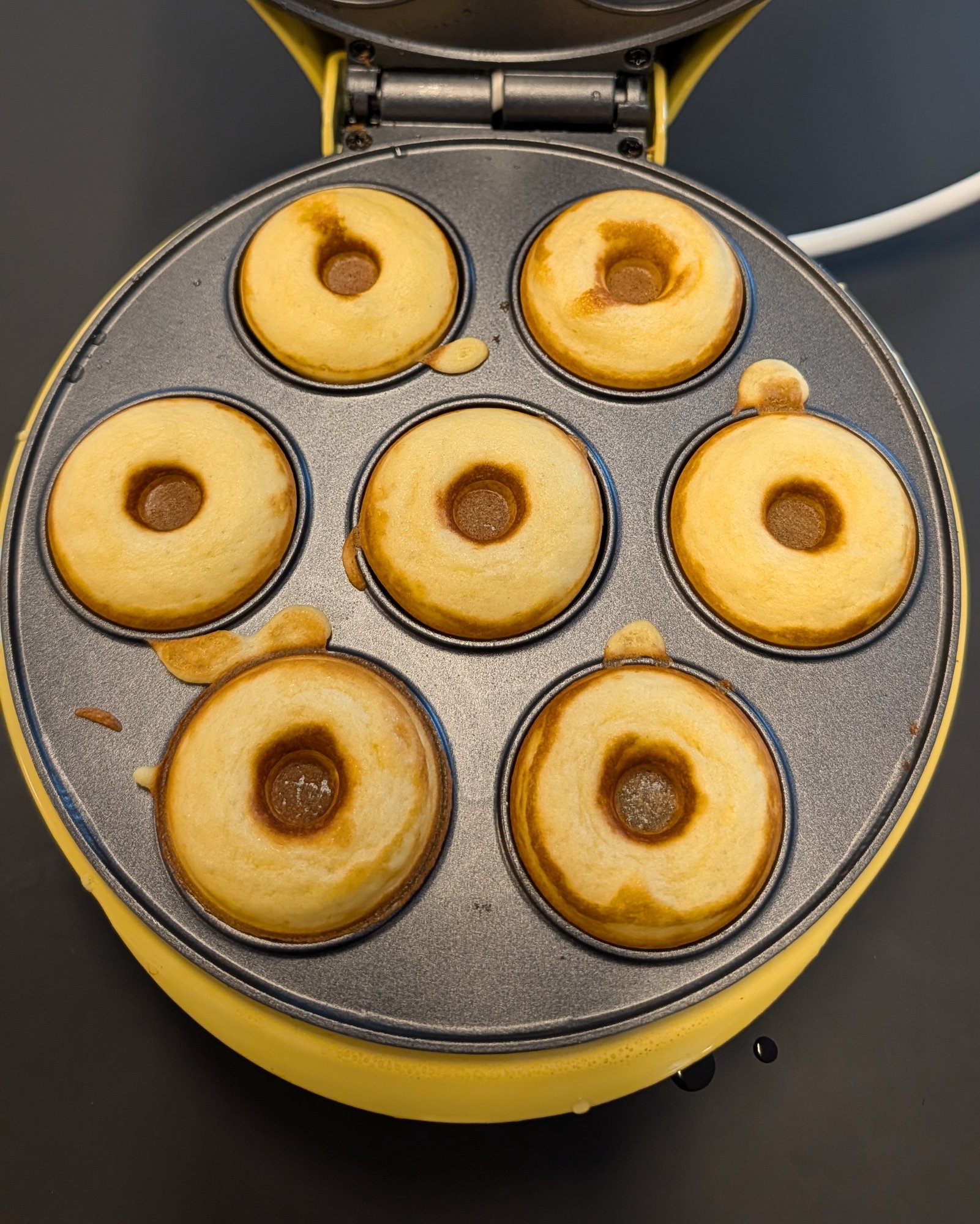 Sieben frisch gebackene goldgelbe Mini-Donuts in den geöffneten Mulden eines gelben elektrischen Donut-Makers, von oben fotografiert