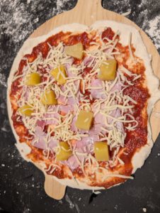 Rohe belegte Pizza auf dem Schieber mit Tomatensoße, Schinken, Ananasstücken und geriebenem Käse