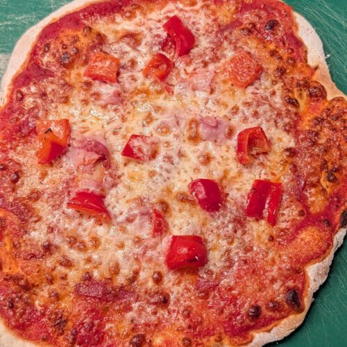 Gebackene glutenfreie Pizza mit Tomatensoße, geschmolzenem Mozzarella, Schinkenstücken und gewürfelter roter Paprika auf einem grünen Brett