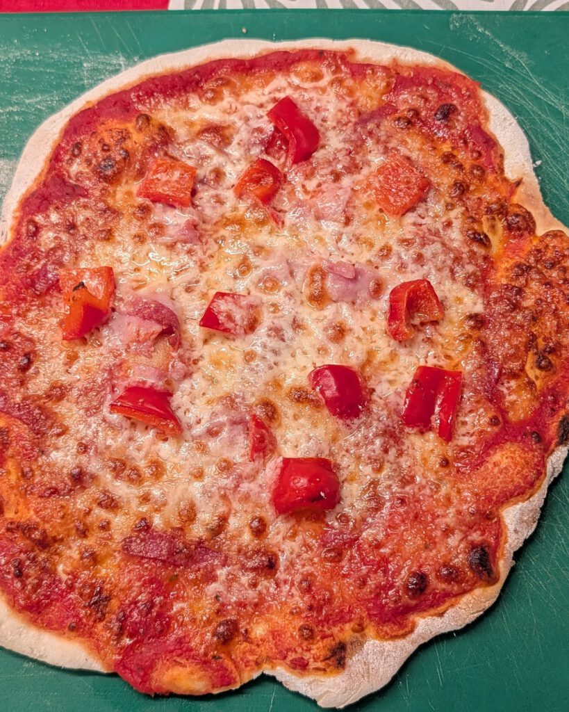 Gebackene glutenfreie Pizza mit Tomatensoße, geschmolzenem Mozzarella, Schinkenstücken und gewürfelter roter Paprika auf einem grünen Brett
