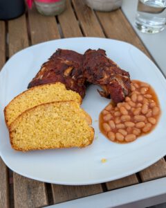 Weißer Teller mit glasierten BBQ-Rippchen, zwei Scheiben goldgelbem Maisbrot und Baked Beans in Tomatensoße auf einem Holztisch im Freien