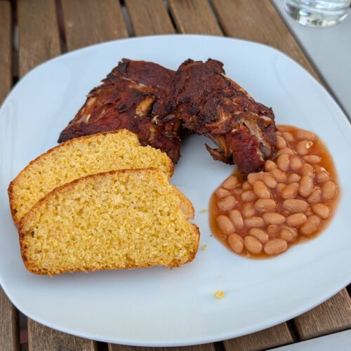 Weißer Teller mit glasierten BBQ-Rippchen, zwei Scheiben goldgelbem Maisbrot und Baked Beans in Tomatensoße auf einem Holztisch im Freien