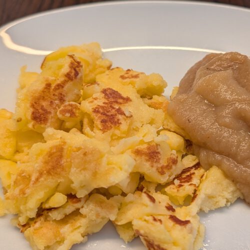 Goldbraune, zerrissene Kaiserschmarrn-Stücke und ein Klecks Apfelmus auf einem weißen Teller auf dunklem Holztisch