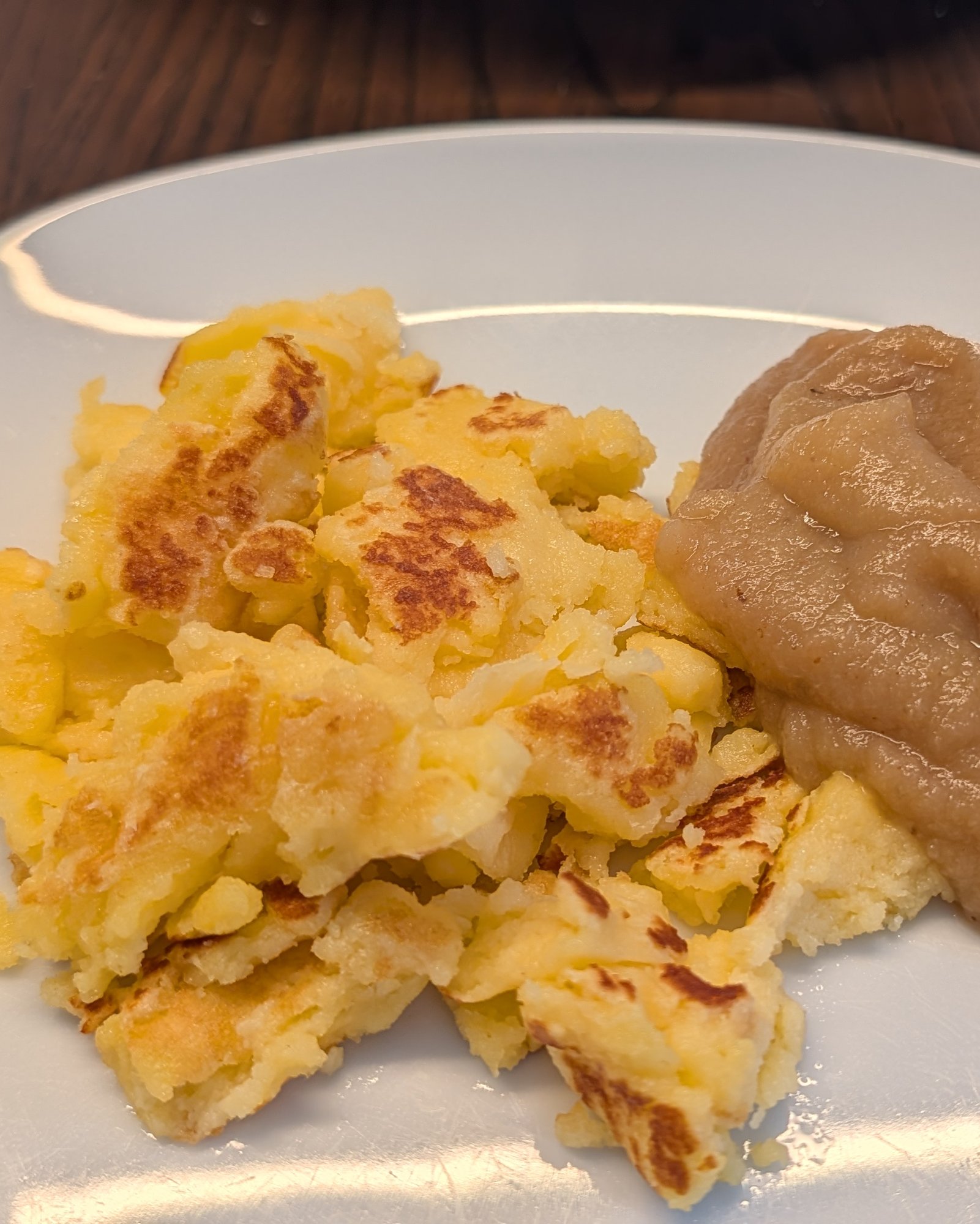 Goldbraune, zerrissene Kaiserschmarrn-Stücke und ein Klecks Apfelmus auf einem weißen Teller auf dunklem Holztisch