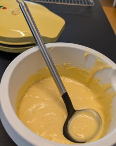 Glatter gelber Donutteig in einer weißen Schüssel mit Schöpflöffel, daneben ein geschlossener gelber Donut-Maker