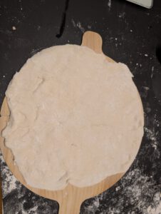 Von Hand flach gedrückter Pizzateig auf einem Holzschieber mit Mehl auf der dunklen Arbeitsfläche