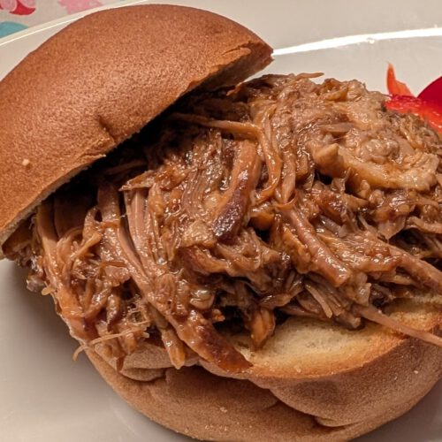 Pulled Pork im glutenfreien Brötchen, daneben rote Paprikastreifen auf einem weißen Teller, dahinter eine Tischdecke mit Herzmuster