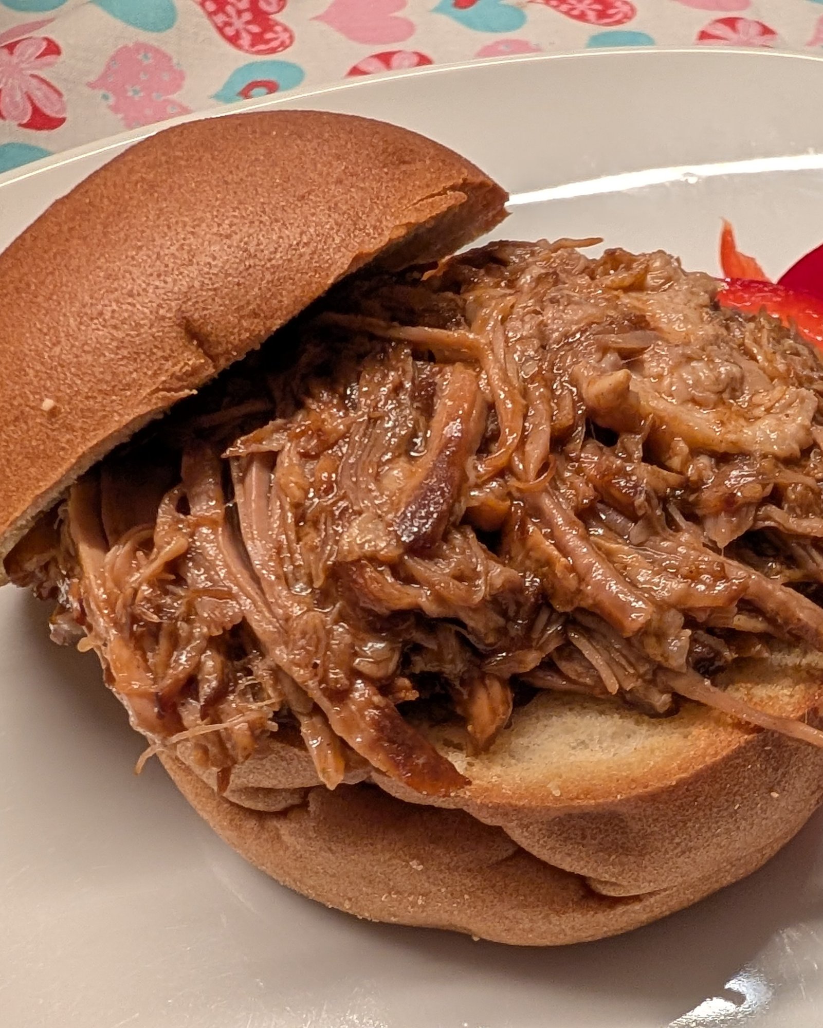 Pulled Pork im glutenfreien Brötchen, daneben rote Paprikastreifen auf einem weißen Teller, dahinter eine Tischdecke mit Herzmuster