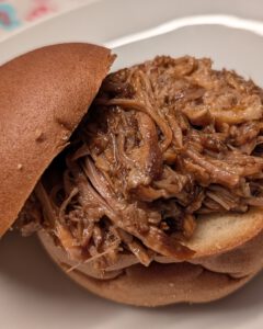 Nahaufnahme eines Pulled-Pork-Burgers im Brötchen mit zur Seite gekippter oberer Hälfte auf einem weißen Teller