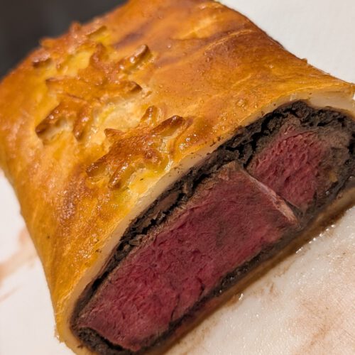 Angeschnittenes Beef Wellington mit rosa Rinderfilet, dunkler Duxelles-Schicht und goldenem Teig mit Schneeflocken-Verzierung auf einem weißen Brett mit grünem Rand