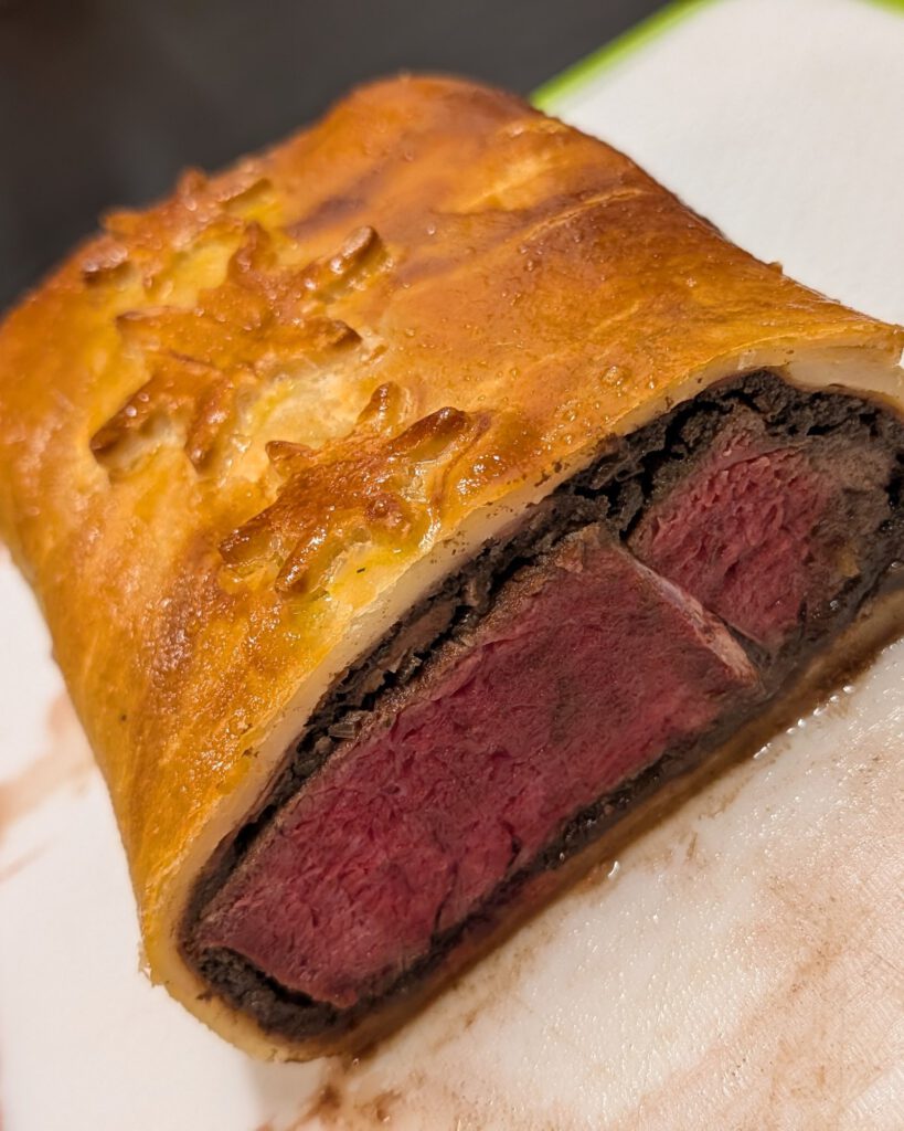 Angeschnittenes Beef Wellington mit rosa Rinderfilet, dunkler Duxelles-Schicht und goldenem Teig mit Schneeflocken-Verzierung auf einem weißen Brett mit grünem Rand