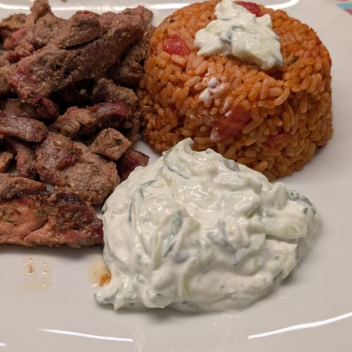 Weiße quadratische Platte mit in Scheiben geschnittenem, angebratenem Gyros, einer Kuppel Tomatenreis mit Zaziki obenauf und einem Klecks Zaziki mit Gurke