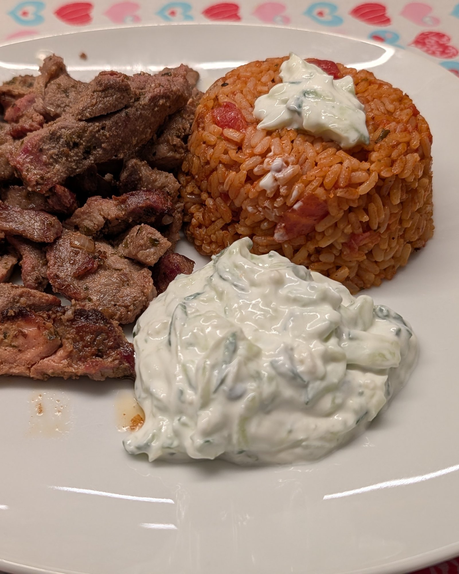Weiße quadratische Platte mit in Scheiben geschnittenem, angebratenem Gyros, einer Kuppel Tomatenreis mit Zaziki obenauf und einem Klecks Zaziki mit Gurke