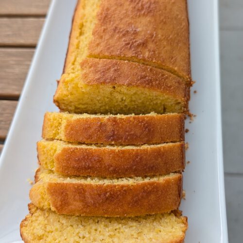 Goldbrauner Maisbrot-Laib auf einer langen weißen Platte, vorn vier abgeschnittene Scheiben mit goldener Kruste und krümeliger gelber Krume