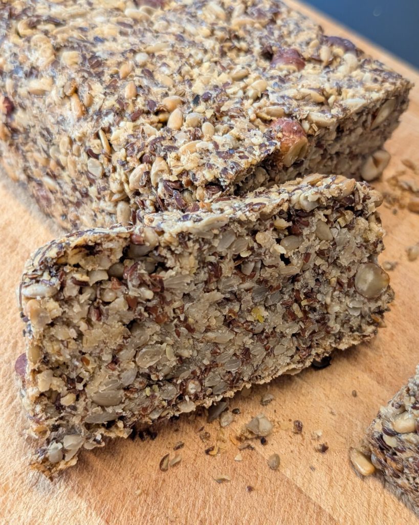 Dichter Saatenbrot-Laib mit einer abgeschnittenen dicken Scheibe davor, die Krume zeigt Sonnenblumenkerne, Leinsamen und ganze Haselnüsse, auf einem Holzbrett