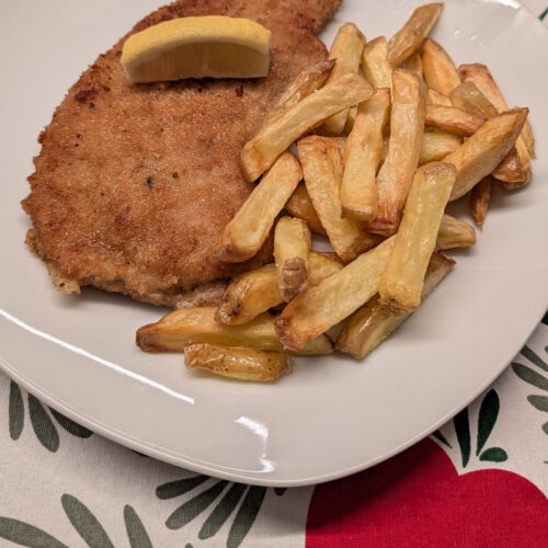 Paniertes Schnitzel mit goldbrauner Kruste auf einem weißen Teller, obenauf ein Zitronenspalt, daneben ein Häufchen goldgelber Pommes frites, auf einer rot-grün-weiß gemusterten Tischdecke