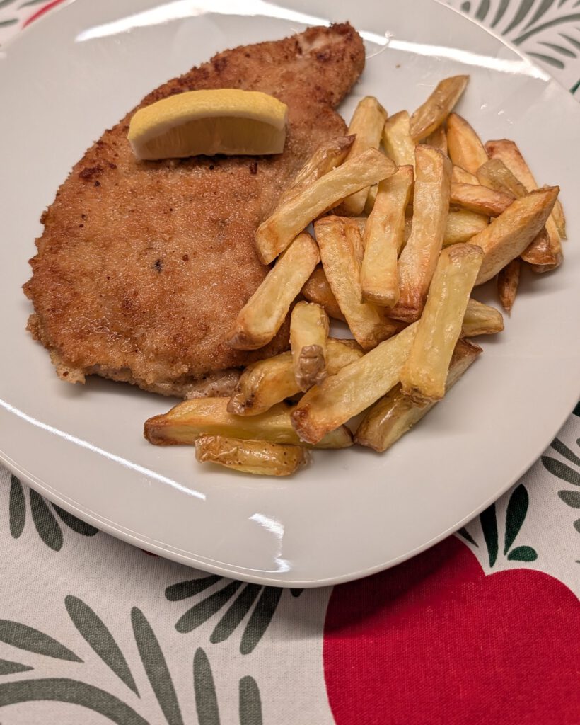 Paniertes Schnitzel mit goldbrauner Kruste auf einem weißen Teller, obenauf ein Zitronenspalt, daneben ein Häufchen goldgelber Pommes frites, auf einer rot-grün-weiß gemusterten Tischdecke