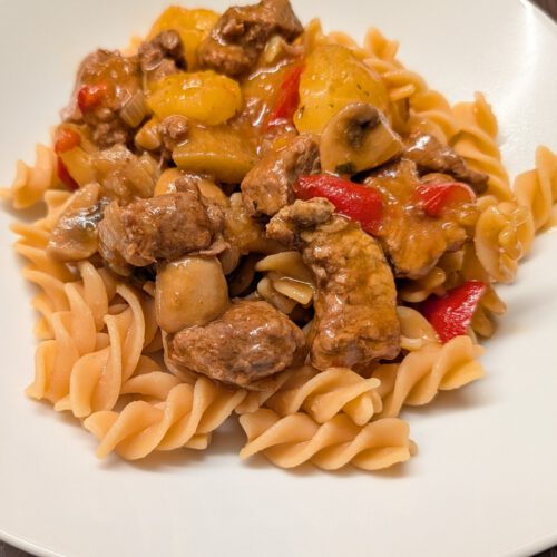 Rindergulasch mit Kartoffelstücken, Champignons und roter Paprika auf Spiralnudeln auf einem weißen Teller auf dunklem Holztisch