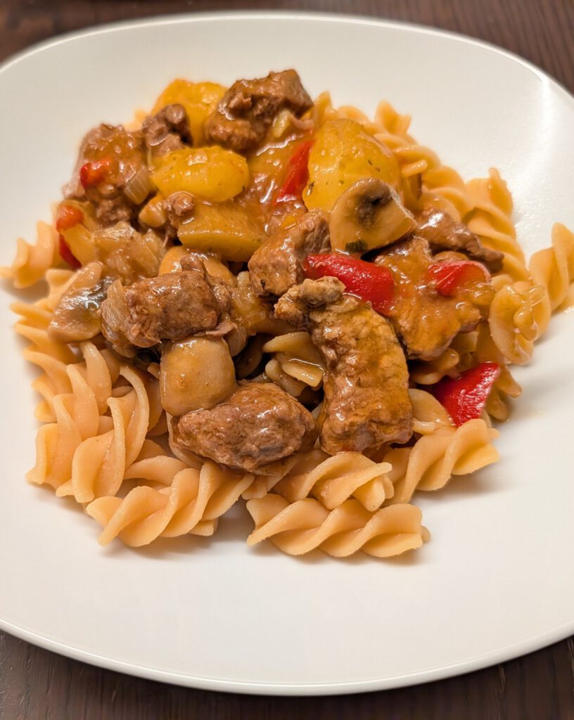 Rindergulasch mit Kartoffelstücken, Champignons und roter Paprika auf Spiralnudeln auf einem weißen Teller auf dunklem Holztisch