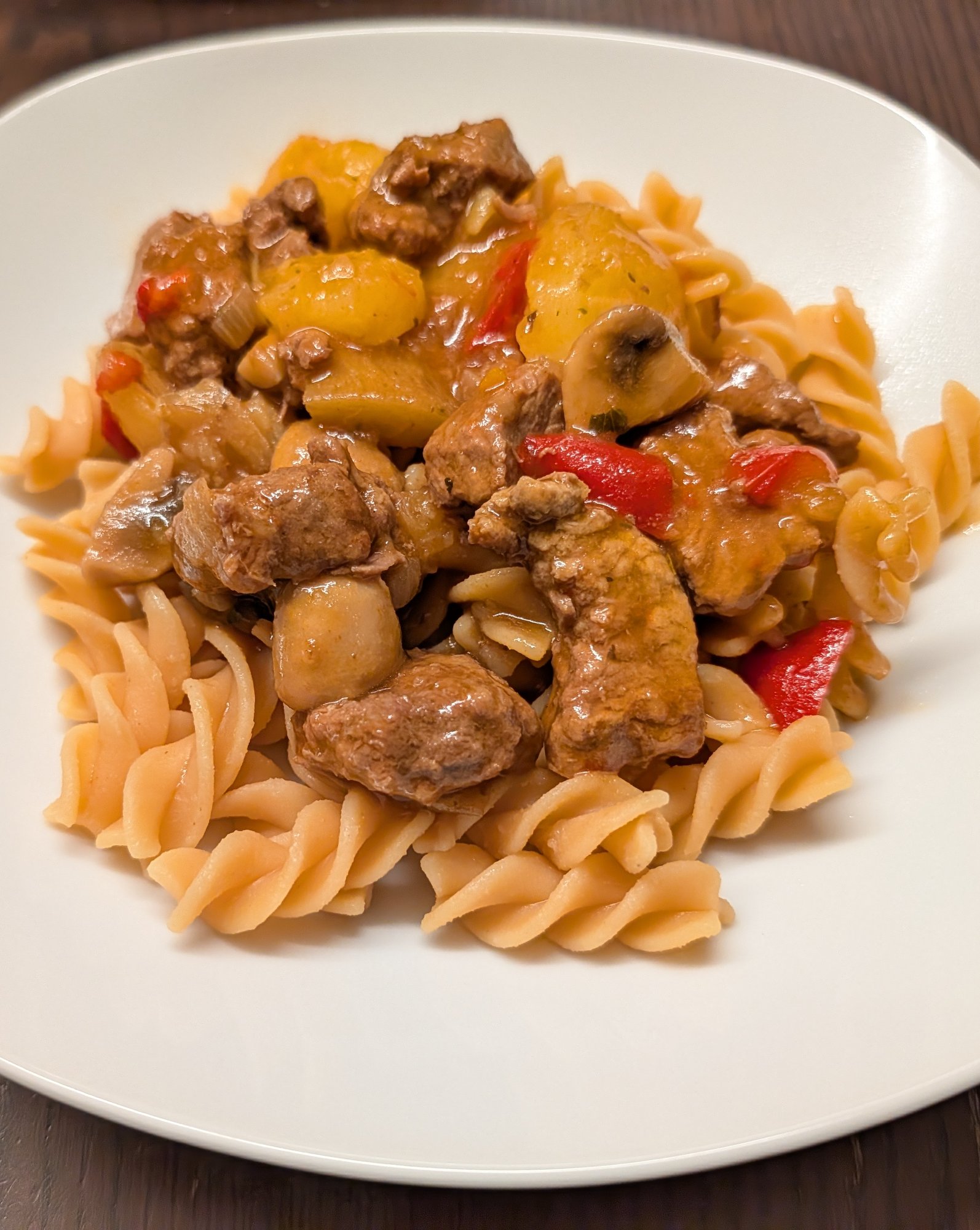 Rindergulasch mit Kartoffelstücken, Champignons und roter Paprika auf Spiralnudeln auf einem weißen Teller auf dunklem Holztisch