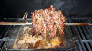 Mariniertes Schweinefleisch dicht auf dem Drehspieß im Gasgrill, darunter eine Glasschale mit Bratensaft
