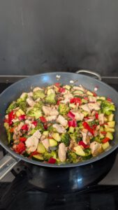 Große Pfanne auf dem Herd mit Hähnchenstücken, Brokkoli, roter Paprika, Zucchini und gebratenen Champignons, gut vermischt