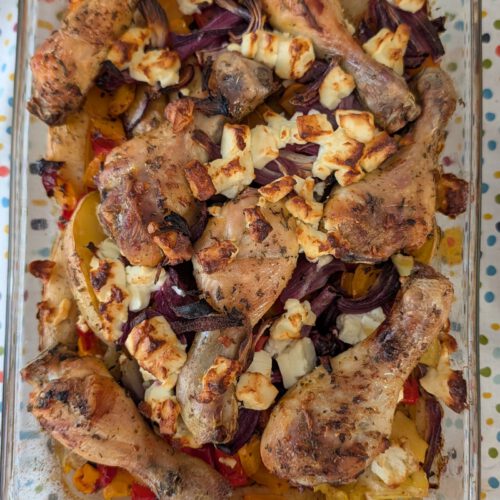 Gebackene Hähnchenschenkel in einer rechteckigen Glasform mit Kartoffelspalten, roter Zwiebel und Paprika, darüber gebräunte Fetabröckchen