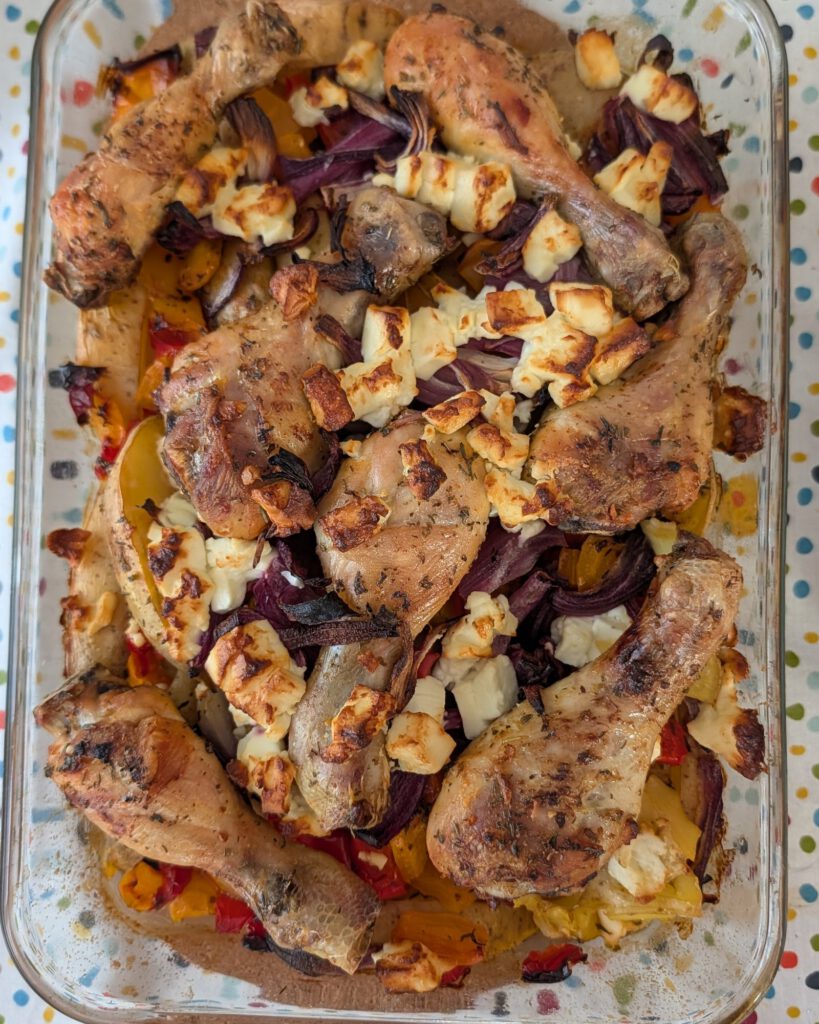 Gebackene Hähnchenschenkel in einer rechteckigen Glasform mit Kartoffelspalten, roter Zwiebel und Paprika, darüber gebräunte Fetabröckchen