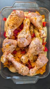 Marinierte rohe Hähnchenschenkel auf Kartoffelscheiben und gewürfelter roter und oranger Paprika in einer rechteckigen Glasform