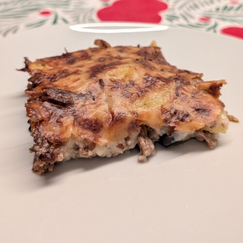Ein Stück gebackene Moussaka auf einem weißen Teller mit gebräunter, dunkel gesprenkelter Oberseite; an der Schnittfläche sieht man Hackfleisch und helle cremige Schichten.