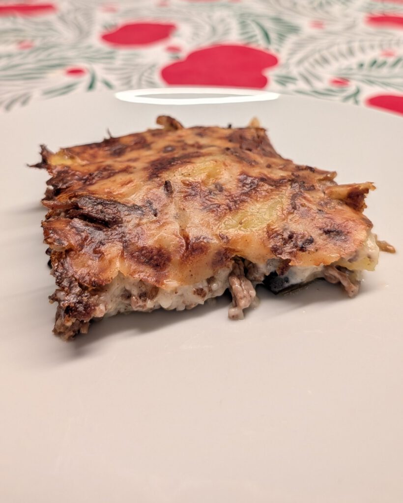 Ein Stück gebackene Moussaka auf einem weißen Teller mit gebräunter, dunkel gesprenkelter Oberseite; an der Schnittfläche sieht man Hackfleisch und helle cremige Schichten.