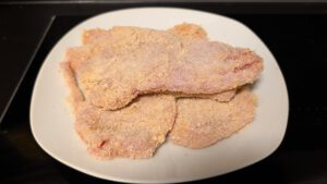 Panierte rohe Schnitzel in hellorangem Paniermehl, auf einem weißen Teller gestapelt, vor dem Ausbacken